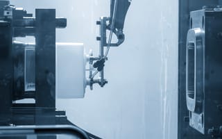 Robotic arm operates a plastic injection molding machine, creating a product in a high-tech, industrial setting. The tone is efficient and precise.