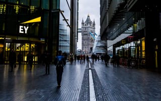 A dimly lit alley between modern buildings leads to a view of the Tower Bridge in London. People walk along the wet pathway, creating a bustling yet serene atmosphere.
