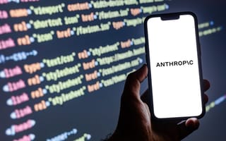 A hand holds a phone with the Anthropic logo on the screen. Lines of code are pictured on a computer monitor in the background.