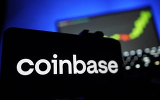 The Coinbase logo is featured on the screen of a smartphone in front of a laptop with a line graph on the monitor.