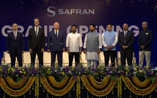 Group of eight men in formal attire stand in front of a stage backdrop with "Safran Grand Opening" text. Garland and floral decorations adorn the stage.