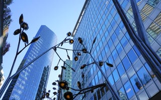 A cluster of modern skyscrapers under a clear blue sky, framed by an artistic metal sculpture with abstract flower shapes in the foreground.