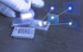 A gloved hand holds a computer chip over other chips on a table, emitting digital connections to futuristic icons, symbolizing advanced technology and innovation.