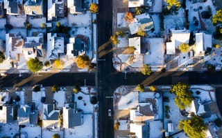 Aerial view of a snowy suburban neighborhood at sunrise, showing a grid of houses with snow-covered roofs and long shadows, creating a tranquil winter scene.
