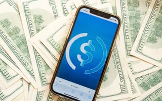 Smartphone displaying a dollar symbol app screen on a pile of hundred-dollar bills, conveying themes of digital finance and currency exchange.