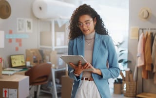 A woman in a blue blazer, standing in a stylish office space, looks at a tablet. Behind her are boxes, a computer, clothes on a rack, and soft lighting.
