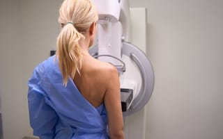 A woman with light blonde hair in a ponytail wears a blue medical gown and stands facing a mammography machine in a neutral-toned room.