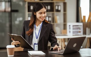 A smiling woman in a blazer works at a desk with a laptop, clipboard, and coffee cup, conveying a professional and productive office setting.