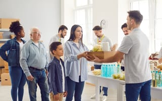A diverse group of people in a well-lit room participate in a food drive. A woman hands a box of groceries to a volunteer. The atmosphere is cooperative.