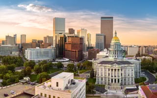 Aerial view of a city skyline during sunset with a majestic white capitol building and golden dome in foreground, surrounded by lush green trees.