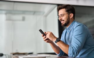 Man in a blue shirt, wearing glasses, sitting at a desk, smiles while looking at his smartphone. A bright office background adds a professional tone.