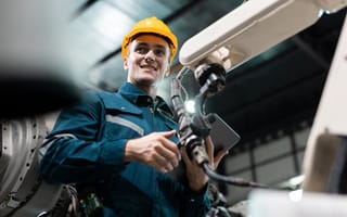 A smiling worker in a yellow hard hat and blue uniform holds a tablet, standing beside industrial machinery in a factory setting.