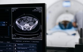 A medical monitor displays a CT scan image in a dim room. In the background, a patient lies on a bed entering a CT scanner, conveying a clinical atmosphere.