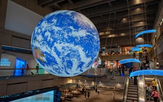 A large illuminated Earth globe hangs in a bustling science museum, surrounded by visitors. The space is modern, with escalators and interactive exhibits.