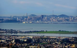 Aerial view of San Francisco skyline and Bay Bridge, with tall buildings, rolling hills, and bay waters under a cloudy blue sky. Urban landscape with greenery.