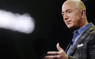 Jeff Bezos wearing a plaid blazer and blue shirt gestures while speaking on stage. The background is dark, creating a focused and serious tone.