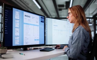 A woman in a denim shirt reviews invoices on dual monitors in a modern office. She is focused and working efficiently, conveying a professional tone.