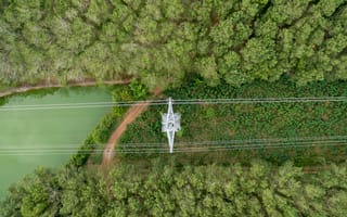 Aerial view of a power line tower intersecting dense green forest, with a winding river on the left. The scene is serene and untouched by human activity.