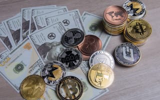 Several hundred dollar bills sit on a table surrounded by gold and silver coins featuring cryptocurrency logos.
