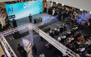 A bird's-eye view of AI Expo 2022, showcasing a large stage with a presenter, seated audience, and vibrant "Robotics for All" signage, exuding an innovative atmosphere.