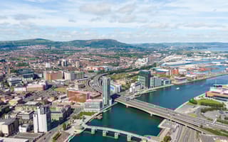 Aerial view of a bustling cityscape featuring modern skyscrapers, bridges over a river, and a distant backdrop of lush green hills under a partly cloudy sky.