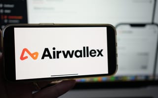 A smartphone screen displays the Airwallex logo with an orange and black design. It is held in front of a blurred laptop screen, suggesting a tech context.