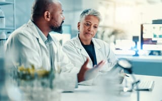 Two scientists in lab coats discuss research in a laboratory setting. One gestures while speaking, and the other listens intently. The atmosphere is focused.