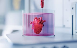 A 3D-printed heart model suspended in pink liquid within a clear container in a lab setting. The scene conveys innovation and scientific research.