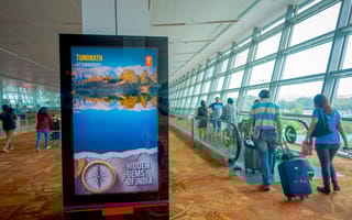 Airport corridor with travelers and luggage on moving walkway. Bright digital screen displays a scenic ad for "Tungnath, Uttarakhand: Hidden Gems of India."