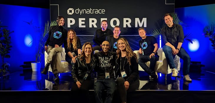 Nine Dynatrace team members sit on a stage with blue backlighting and a sign reading “Dynatrace Perform.”