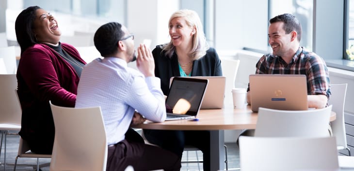 Photo of four colleagues sitting at a table with laptops open, laughing at a joke.