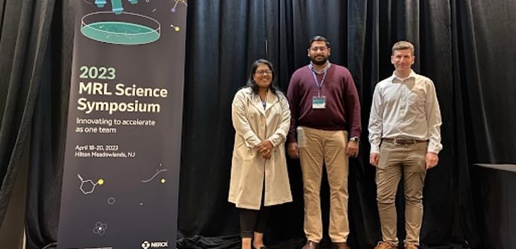 Three Merck team members stand next to banner for 2023 MRL Science Symposium