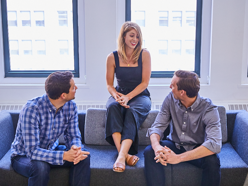 Erin Lowrey sits on the top of a sofa surrounded by captivated coworkers