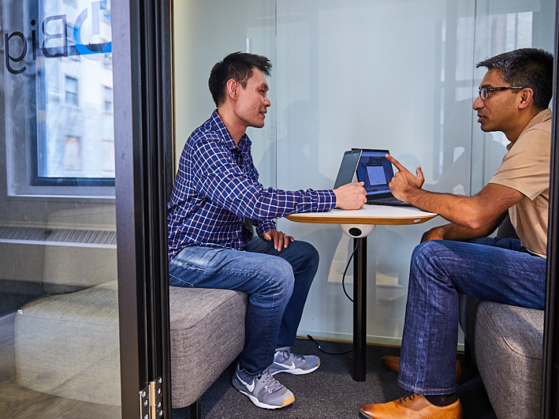 two Trade Desk employees sit facing each other while holding laptopsl