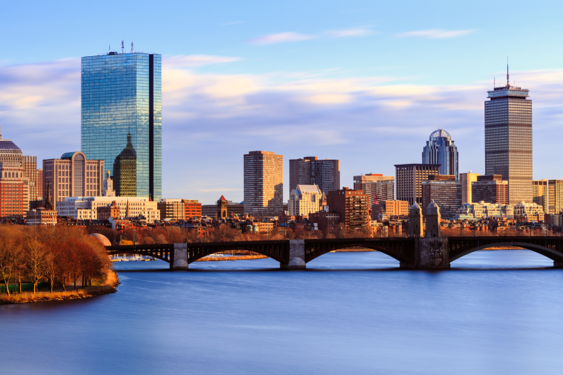 A landscape view of the Boston skyline and harbor. The sunset reflects off the buildings to the west.