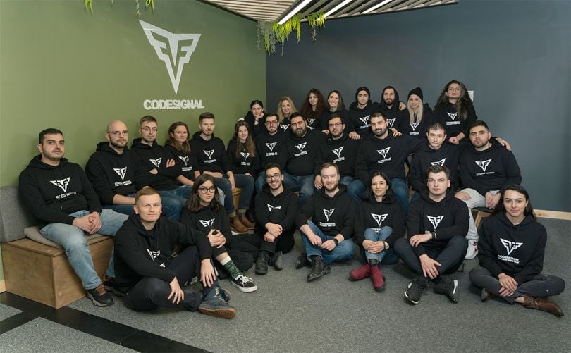 CodeSignal group photo in the office with company logo on the wall behind them
