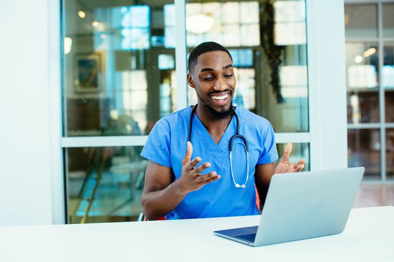 male doctor wearing blue scrubs uniform using laptop to talk to patient online