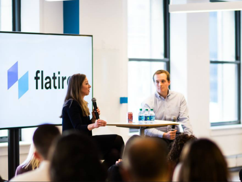 A professional man and woman speak to a room of people