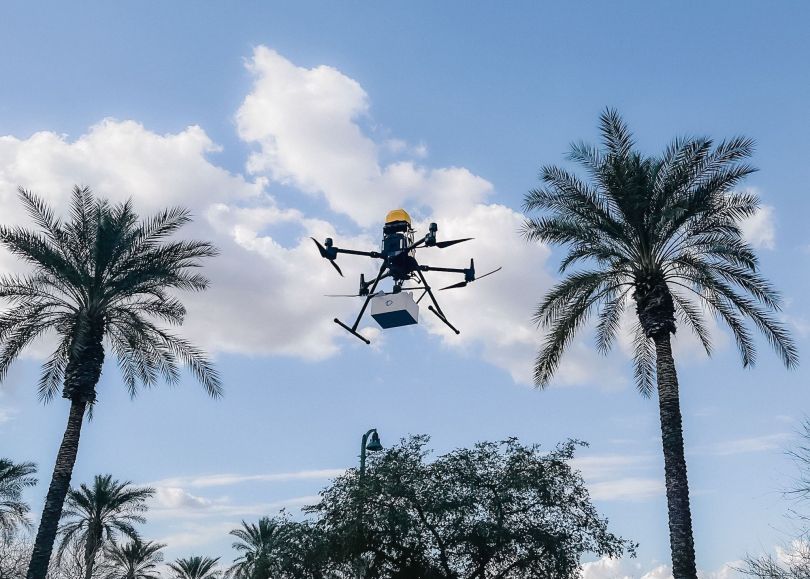 A drone holding a package hovers against the clear sky.