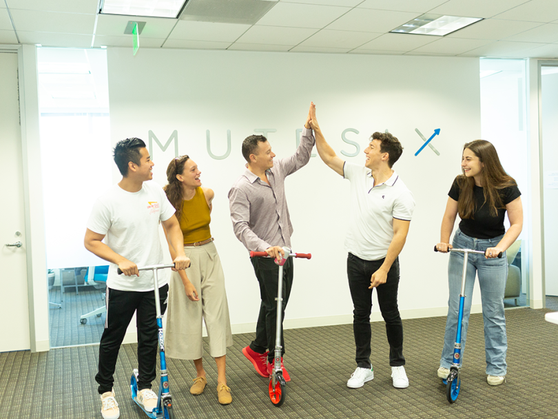 MuteSix team high fives on scooters below the MuteSix logo