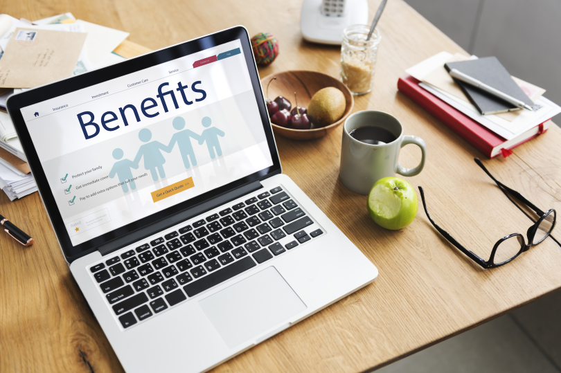 laptop on a desk with a pair of glasses and a cup of coffee. A benefits portal appears on the screen. 