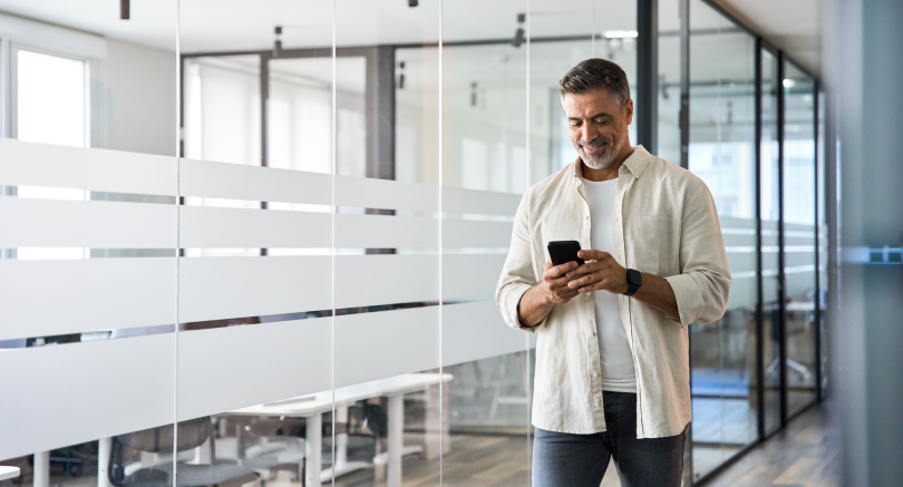middle-aged man with salt and pepper hair dressed casually in jeans and a t-shirt with button-up shirt open in the front. He looks at his cell phone while smiling and walking in an office. 