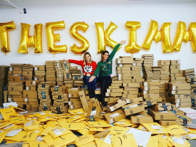 Two women sit in front of gold balloons that spell out The Skimm