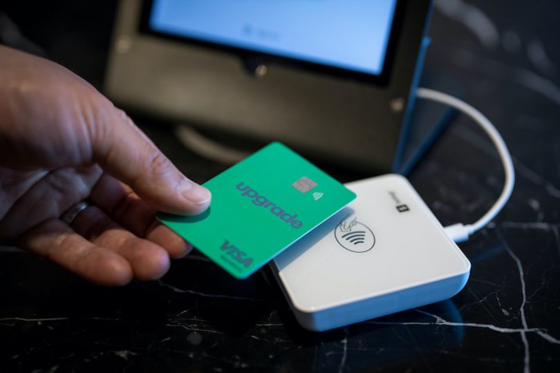 A hand holding an Upgrade-branded Visa card over a touch payment machine, as if making a purchase.