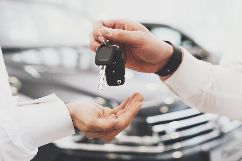 A person is depicted handing car keys to someone in a dealership.