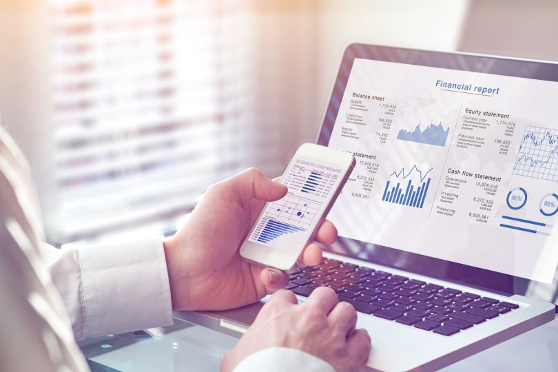 Person looking at financial data on their smartphone. There is a data dashboard pulled up on a laptop in the background. 