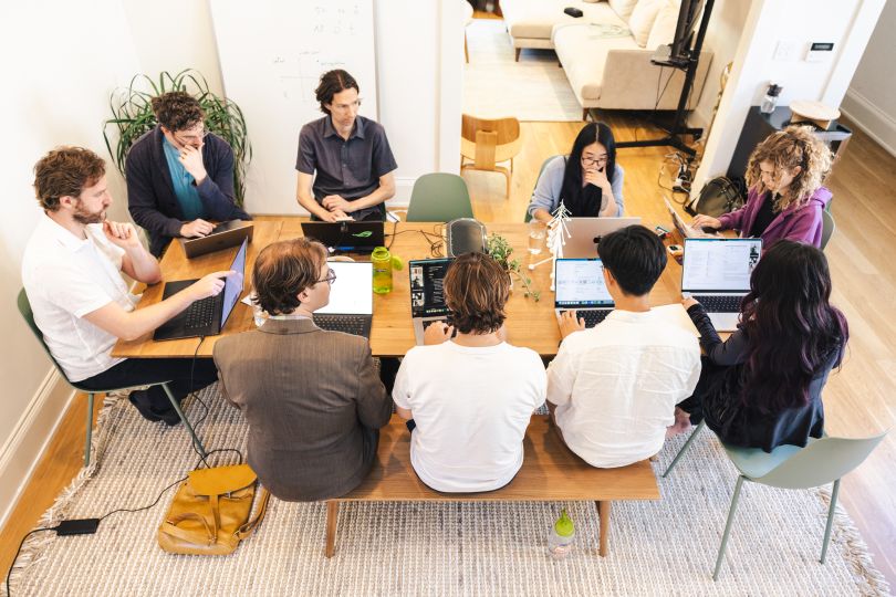 A group of people sit at a dining table working together on laptops.&nbsp;