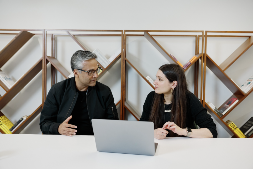 Two people sit at a table in front of an angled bookshelf, having a discussion.&nbsp;