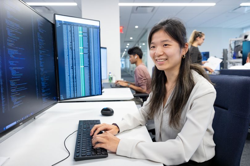 Citadel Software Engineer Cindy Zhang sits at a computer with two monitors, hands over the keyboard, smiling. 