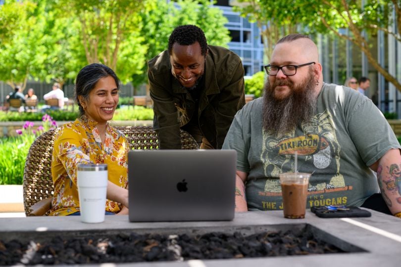 Three Cox Enterprises employees look at information on a laptop screen while working together in an outdoor space at one of the company's offices